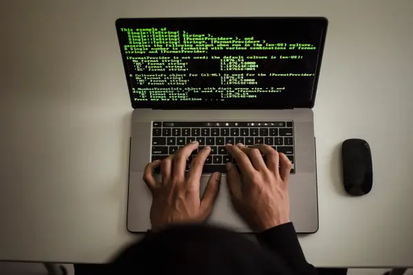 Uma vista superior de um notebook com código verde em tela, com mãos digitando no teclado.